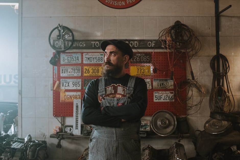 Portrait of a bearded mechanic in a vintage garage surrounded by car parts and license plates.